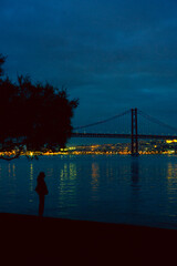 Lisbon April 25 bridge on a cloudy day blue hour, a person and tree silhouette lights on