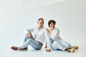 Middle aged couple wearing white shirts and jeans sitting on grey background and looking away
