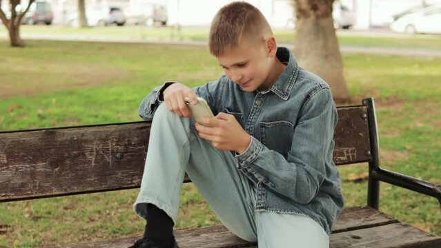 camera captures slow-motion footage circling around a European-looking boy sitting on a bench in urban park, watching a funny video on his phone and smiling