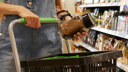 Close-up of a young woman's hands putting a glass jar of instant coffee into a shopping cart