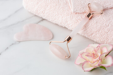 Rose quartz facial roller and gua sha stone for anti-aging skin care massage with pink rose flower and towel on while marble table.