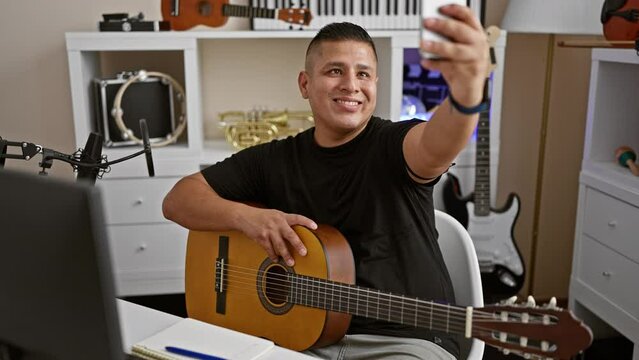 Smiling young latin man, an artist in his prime, striking a melody on his classical guitar, takes a selfie with his smartphone in a music studio