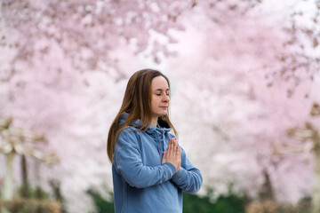 A young woman is engaged in breathing yoga practice against the backdrop of a spring blossoming sakura alley. Hands folded in a namaste gesture. Concept of mental, physical health and balance.