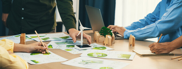 Windmill model represented renewable energy and wooden block represented eco city was placed on green business meeting table with environmental document scatter around. Front view. Delineation. - obrazy, fototapety, plakaty