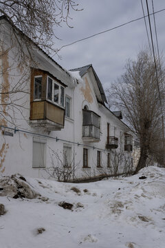 The peeling facade of an old house. The inscription on the facade- 27 Lenina Street. Drifts of dirty snow, a tree, power lines. A provincial town in northern Russia.