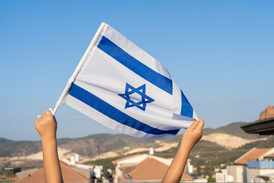 Children's Hands Hold Israeli Flag Over Sunny Rooftop Sky.