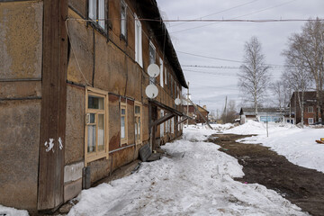 An old house in a provincial town in Russia. Peeling facade, old windows, satellite TV, thawed snow. Greay sky. In the distance, a banner of an electrical equipment store.