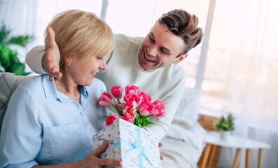 Sincerely happy mature woman looks on tulips bouquet and gift box from her young son while sitting on the couch at home. Mothers day, birthday or womens day concept - 771004192