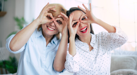 Emotional and excited laughing mother and daughter grimacing funny faces and showing glasses with help of hands and having fun - 771004316