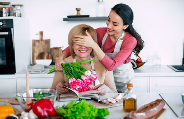 Surprise in Mother's day on domestic kitchen. Excited beautiful daughter covering mother's eyes while making greeting her with tulips in hand. Women day gifts - 771004345