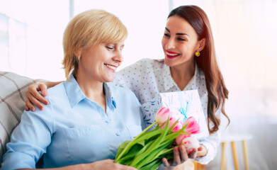 Beautiful smiling young brunette woman greeting and congratulating her mother with flowers and gift while they sitting on the couch at home. Mothers day or womens day concept - 771004361