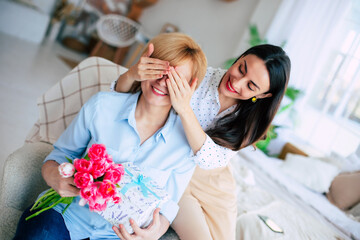 Family surprise. Cute female covering mother's eyes and giving her present and gives a bouquet of flowers to tulips at home. Mothers day, birthday or womens day concept - 771004389