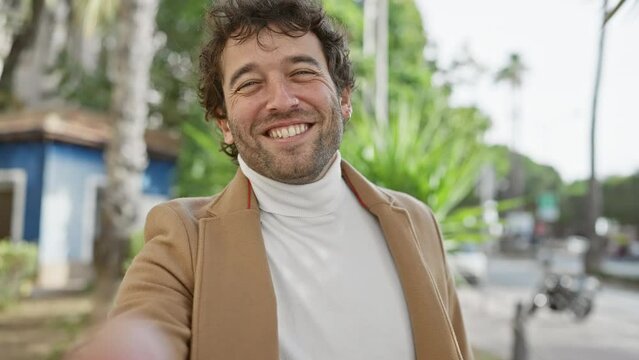 A smiling young hispanic man in a coat standing on a city street extends his hand towards the camera, giving a sense of welcoming and friendliness.