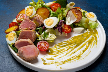 Delicious Nicoise salad with fried tuna, vegetables, lettuce and egg in a plate on a concrete background