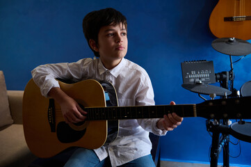 Serious adolescent boy perform sound melody, singing song while playing acoustic guitar in modern...