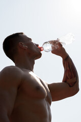 A shirtless athletic man with a tattoo on his arm hydrates by drinking water from a clear bottle against a bright sky backdrop. The image conveys concepts of fitness, health, and hydration.