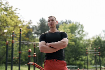 A fit, muscular athlete in sportswear stands confidently with arms crossed in an outdoor park with workout equipment. The scene captures the essence of strength, determination, and healthy lifestyle.