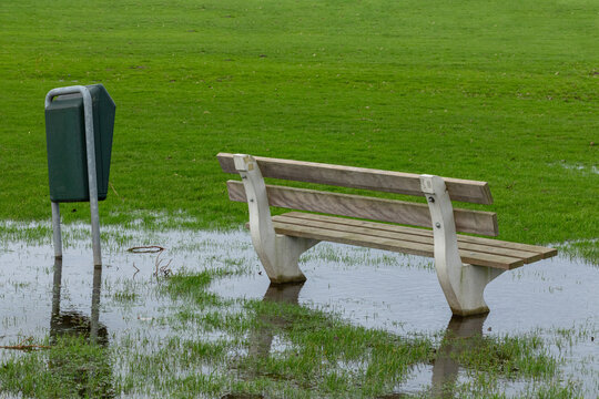 A public bench and trash can in the grass surrounded by water from abundant heavy rain in a park.