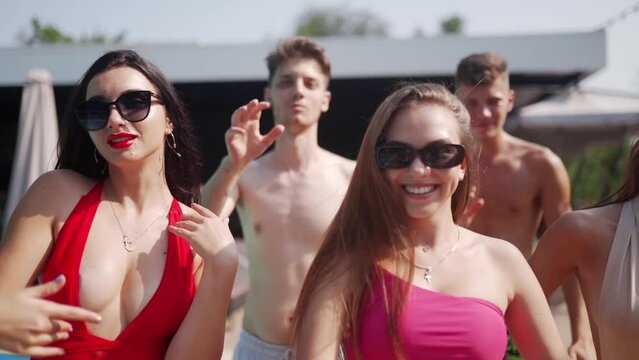Teenagers models have rest on holidays near swimming pool in swimwear. Couples flirting and sending air kisses on summer club party, dancing, waving and laughing with friends in bikinis.