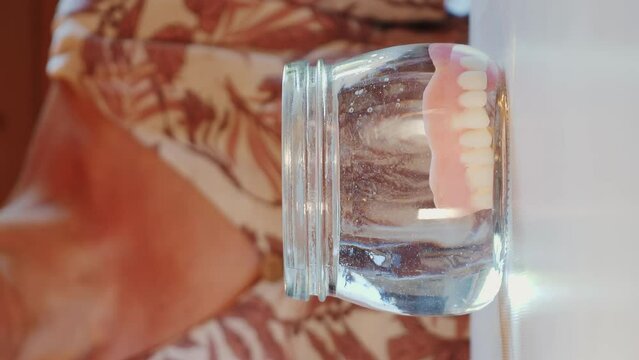 Woman putting false teeth dentures in glass of water. Vertical video