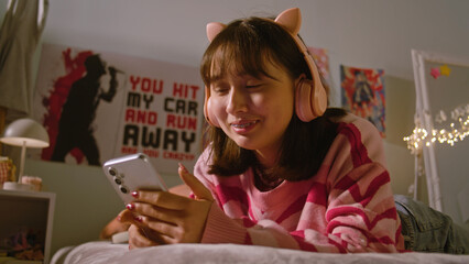 Fototapeta na wymiar Teenage girl listens to music in headphones and scrolls social media using mobile phone lying on the bed in the cozy bedroom. Asian girl spending pastime and having fun at home. Lifestyle concept.
