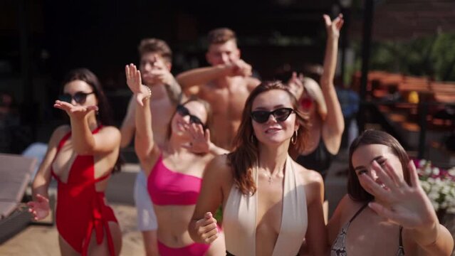 Group of friends in colorful bikinis walking at swimming pool in luxury club outdoor terrace on summer party. Female and male in swimsuits going to chill out on vacation in beach resort.