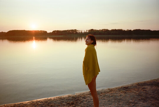 Cheerful woman in  bath towel standing on lakeshore at sunset 