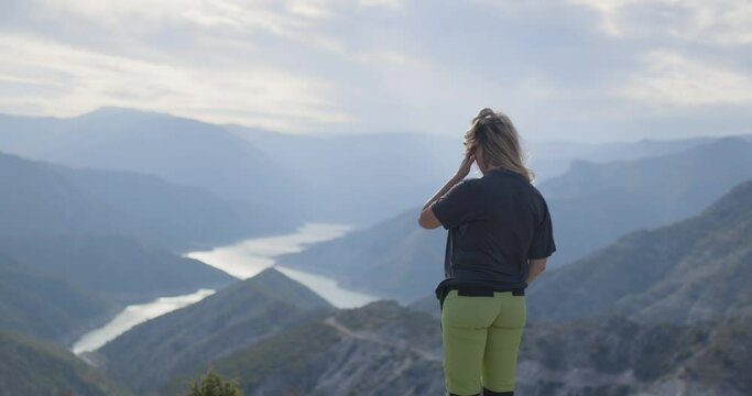 Hiker girl enjoying a view of river lake from high in the mountains. Standing on a cloudy and windy weather.