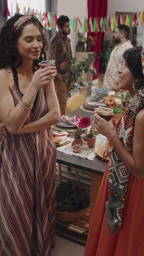 Medium vertical footage of two young beautiful Mexican women in bright head scarves, strappy dresses chatting at Cinco de Mayo party, holding margarita cocktails, then saying toast and smiling