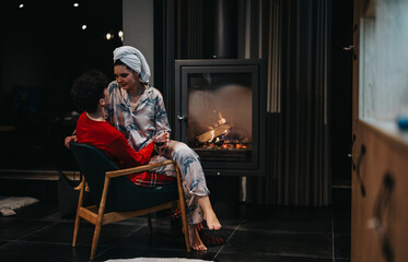 Intimate and warm scene of a couple in pajamas sharing a moment with a glass of wine near a glowing...