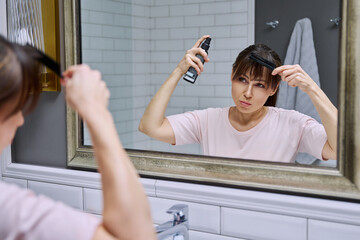 Middle-aged woman with setting spray for styling hair fixation comb looking in mirror