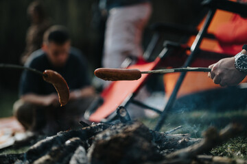 Close-up of sausages cooking on sticks over campfire with friends in the background enjoying outdoor camping fun - obrazy, fototapety, plakaty