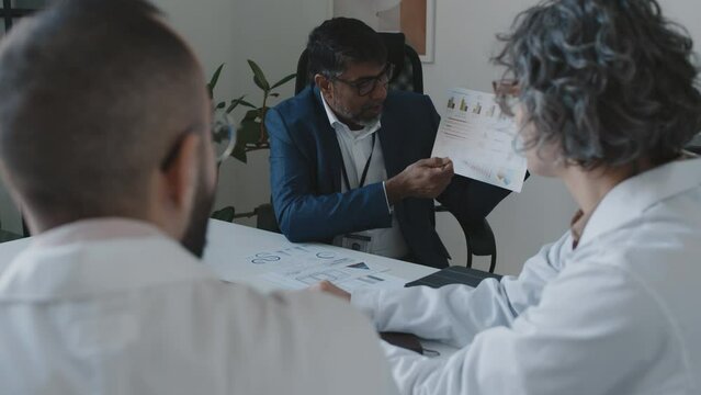 Medium over-shoulder shot of pushy Asian male hospital executive sitting in office with medical director and chief surgeon, suggesting new ways to increase revenue, doctors disagreeing, shaking heads