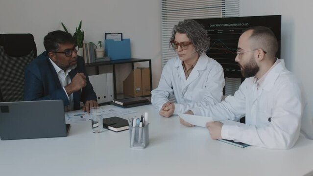 Medium shot of Indian male hospital executive sitting at table in office with diverse medical department heads, looking at financial performance graphs, discussing reasons for decrease in revenue