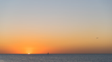 Silhouette of ship on the sea horizon and an airplane flying in the sky above it during beautiful and calm sunset.