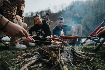 A group of friends gather around a campfire, cooking sausages on sticks, enjoying nature and good...