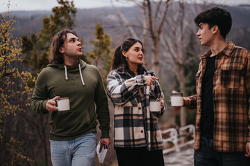 Three friends chatting and holding coffee mugs while standing outdoors, enjoying a relaxing time...