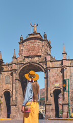 Fototapeta na wymiar Young woman traveler in brown hat walks through of the Santa Clara Arch in San Francisco square. Cusco, Peru.