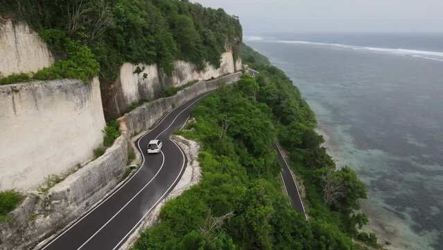 Aerial view, a white car driving along the cliff-side asphalt road and the sea located on Tanah Barak beach, Bali island, Indonesia.