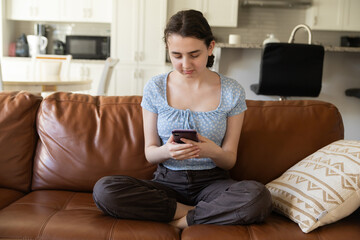 Teenage Girl Texting on Smartphone at Home. A young girl sits comfortably on a brown leather couch,...