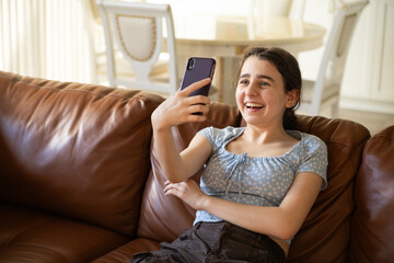 Smiling Teen with Smartphone on Sofa.  Joyful teenage girl lounging on a couch, happily using her...