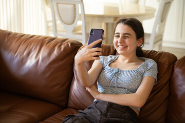 Smiling Teen with Smartphone on Sofa.  Joyful teenage girl lounging on a couch, happily using her...