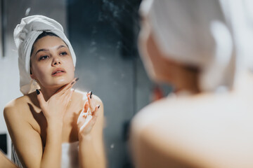 A young woman with a towel on her head applies skincare products while looking into a bathroom...