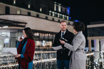 Three young people engage in a business discussion at night, with city lights and holiday decorations in the background.