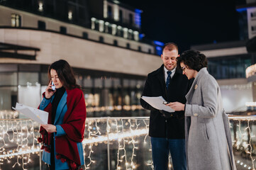Naklejka premium Three professionals gathered outdoors at night during winter, discussing papers with festive lights in the background.