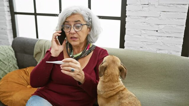Senior woman with glasses and grey hair sitting on couch talking on phone and holding thermometer next to her brown dog in a cozy living room