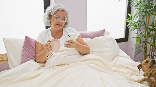 Mature woman in glasses holding cup and phone with dog on bed indoors.