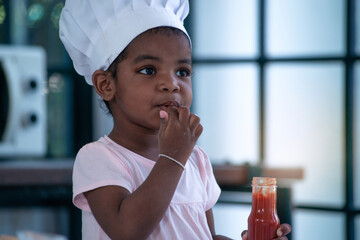 Little African girl wearing a chef's hat in the kitchen, she tastes ketchup in a bottle