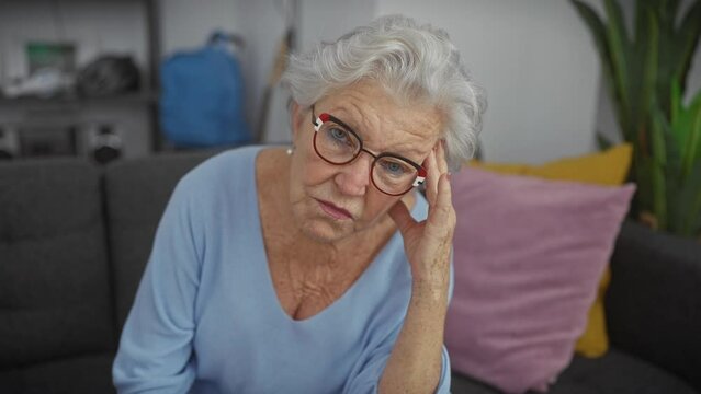 A pensive elderly woman with grey hair and glasses sits at home, concern evident in her expression.