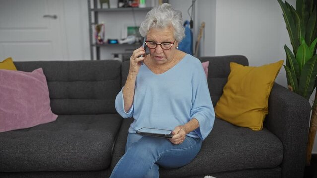 Elderly woman in blue seated on sofa using smartphone and tablet in cozy living room